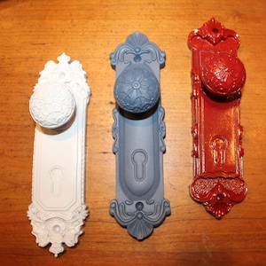 May include: Three ornate door knobs in white, gray, and red. Each knob has a decorative backplate with a keyhole and floral detailing. The knobs are made of metal and are likely for interior doors.