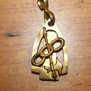 May include: A gold-colored metal pendant in the shape of a pointed oval with a decorative design of a chain, spear, and axe inside.