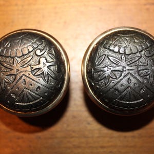May include: Two antique, round cabinet knobs with a dark, textured finish and intricate floral and geometric patterns. Each knob has a gold-colored rim. The knobs are displayed on a wooden surface.