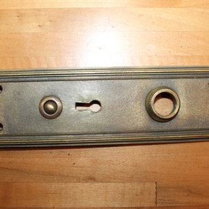 May include: A brass door plate with a keyhole, a round knob, and two smaller round knobs. The plate has a decorative border.