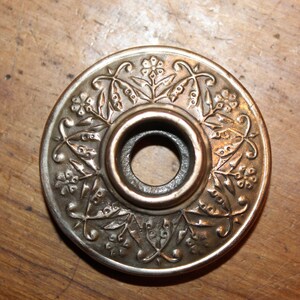 May include: A round, silver-toned metal door knob with a floral design. The knob has a central hole for a key.