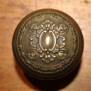 May include: A round, antique-style brass door knob featuring a central oval medallion surrounded by intricate embossed floral and leaf motifs, enclosed within a decorative circular border with a repeating scalloped pattern.