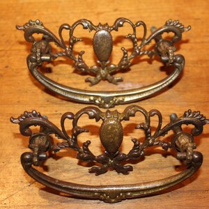 May include: Two antique bronze drawer pulls with ornate, scrolling designs. Each pull features a central oval element and curved handle. The hardware is set against a wooden surface.