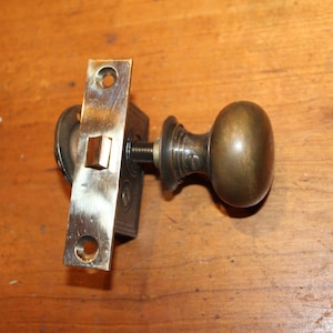 May include: An antique brass door knob and latch set. The knob is oval-shaped, and the latch plate is rectangular with screw holes. The metal has a tarnished appearance, suggesting age. The set is designed for a door.