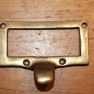 May include: A rectangular brass drawer pull with a handle and four screw holes. The pull has a recessed area for a label. The brass has a slightly aged appearance, suggesting vintage or antique origins.