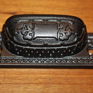 May include: Black cast iron drawer pull with an ornate design. The pull has a rectangular base with a rounded top and two holes for mounting.