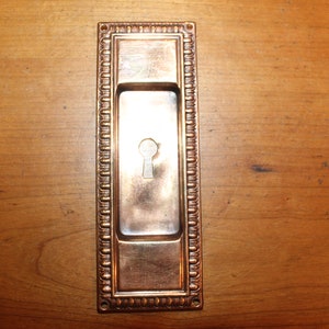 May include: A rectangular brass door plate with a decorative border and a keyhole in the center.