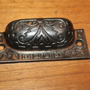 May include: A black metal drawer pull with an ornate design. The pull has a rectangular base with a rounded top and a decorative floral pattern.