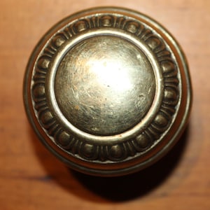 May include: Close-up of a vintage brass doorknob. The doorknob has a circular design with a central disc and an outer ring with a repeating pattern. The brass has a slightly aged appearance.