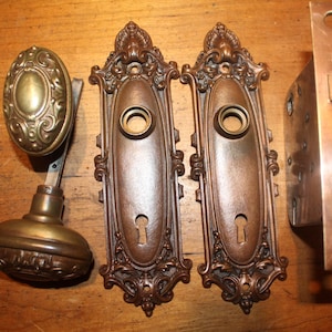 May include: Antique door hardware set featuring a brass doorknob and two ornate bronze-colored backplates. The set includes a matching door latch. The hardware has an aged patina, suggesting vintage or historical use.