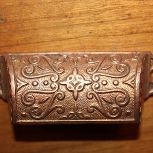 May include: A vintage, ornate, copper-toned drawer pull with a decorative, swirling design.