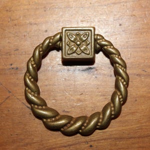 May include: A gold-toned metal ring pull with a decorative, twisted rope design. The ring pull has a square, ornate, floral-shaped center piece.