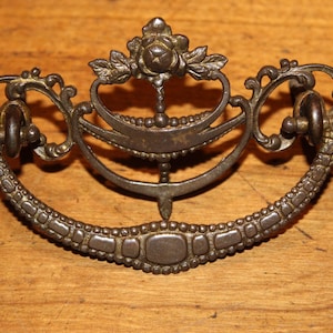 May include: Antique bronze-toned drawer pull with ornate floral and scrollwork details. The handle features a central rose design, curved handle, and decorative beaded trim. A vintage-style hardware piece.
