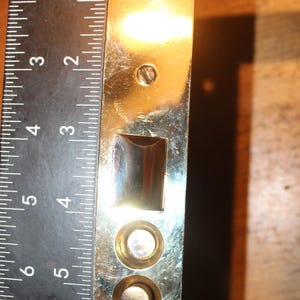 May include: A shiny, rectangular metal door plate with a ruler alongside it. The plate has several holes and a rectangular cutout. The ruler shows measurements in inches. The plate is approximately 8 inches long.