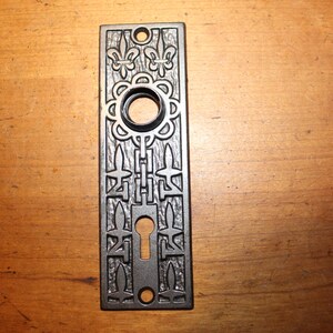 May include: A rectangular, dark gray metal door plate with ornate floral and geometric designs. It features a keyhole, a circular opening for a doorknob, and decorative elements including fleur-de-lis motifs. The plate is likely for a vintage or antique door.