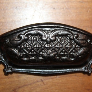 May include: Black cast iron drawer pull with an ornate design. The pull has a curved shape and a decorative pattern of swirls and diamonds.