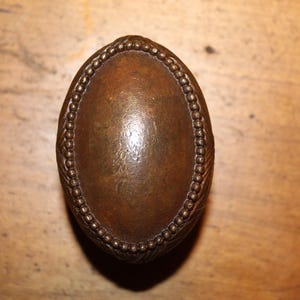May include: Oval-shaped, antique bronze door knob with a beaded border. The center of the knob has a smooth, polished surface, while the border features a detailed, textured design. The knob is likely made of metal.