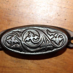 May include: A black cast iron drawer pull with an ornate floral design. The pull is oval shaped and has two holes for mounting.