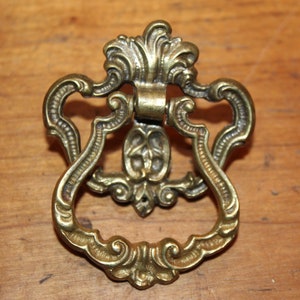 May include: Ornate brass drawer pull with a decorative floral design. The pull has a ring-shaped handle and a detailed, intricate design.