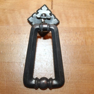 May include: A silver-toned door knocker with a decorative, diamond-shaped backplate. The knocker is shaped like a rectangle with rounded corners and a ring in the center.
