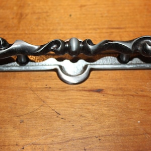 May include: A black metal drawer pull with a decorative scroll design and a rectangular base.
