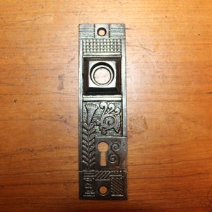 May include: A vintage cast iron door plate with an intricate design featuring a keyhole and floral motifs. The plate is black and has a worn, antique finish.