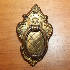 May include: A gold-toned metal cabinet pull with an ornate design and a ring handle. The pull has a diamond-shaped pattern on the front.
