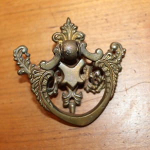 May include: Ornate brass drawer pull with a decorative, scrolled design. The pull has a ring handle and a central, star-shaped accent.