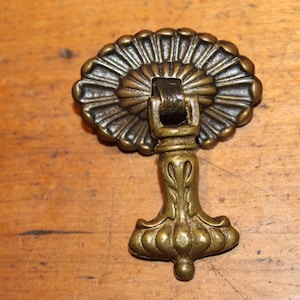 May include: Antique brass drawer pull with a decorative, floral design. The pull features a detailed, fan-shaped backplate and a stylized stem. The hardware is likely for a cabinet or drawer, adding a vintage touch to furniture.