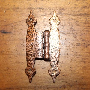 May include: A decorative bronze-colored hinge with a textured surface. The hinge has a central barrel and two ornate, elongated plates with pointed finials and heart-shaped cutouts. The hinge is designed for furniture or cabinetry.