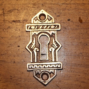 May include: Ornate, antique-style keyhole cover in a bronze finish. The cover features a keyhole, decorative geometric patterns, and two circular holes. The keyhole cover is likely made of metal and is set against a wooden surface.
