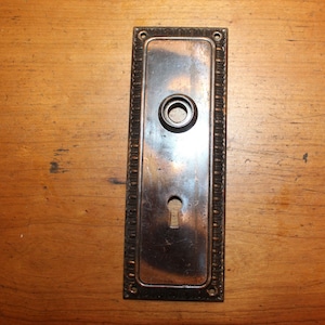 May include: A vintage brass door knob with a keyhole and a decorative border. The knob is mounted on a rectangular plate.