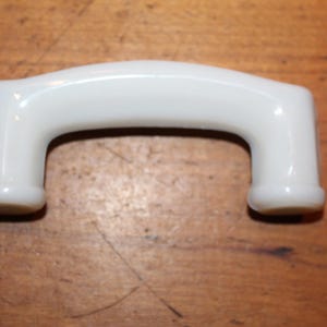 May include: White porcelain cabinet door pull handle. The handle is shaped like a rounded arch with two rounded ends.