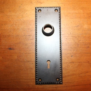 May include: A black metal door plate with a keyhole and a round hole for a knob. The plate has a decorative border.