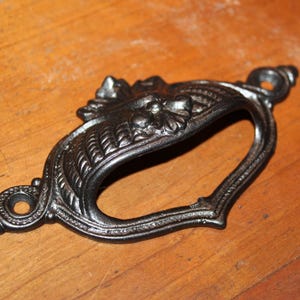 May include: A black metal drawer pull with an ornate design. The pull has a decorative floral pattern and a curved shape.