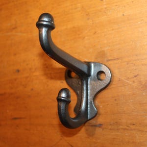 May include: A black cast iron coat hook with a double hook design. The hook is mounted on a decorative backplate with two screw holes. The hook is shown against a wooden background.