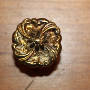May include: A gold-toned metal drawer pull with a floral design. The pull has a raised, ornate design with a central flower motif.