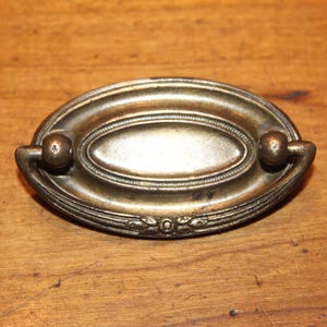 May include: Oval-shaped antique brass drawer pull with a central oval design and decorative detailing. The pull features two rounded handles and is set against a wooden surface. A vintage hardware piece.