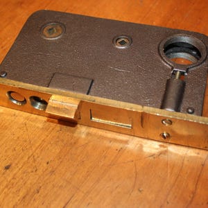 May include: A vintage door lock with a brass-colored faceplate and a dark brown metal body. The lock features a keyhole, a latch, and a handle. The hardware is likely for a door or cabinet.