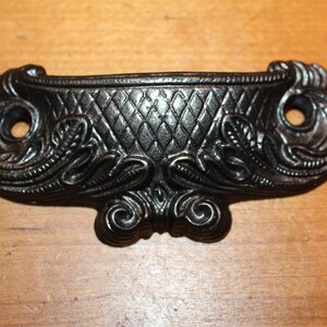 May include: Black metal drawer pull with a decorative, ornate design. The pull has a diamond pattern in the center and a scroll design on the sides.