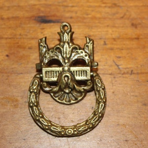 May include: A gold-toned metal door pull with a decorative design. The pull has a ring handle and a detailed, ornate design.