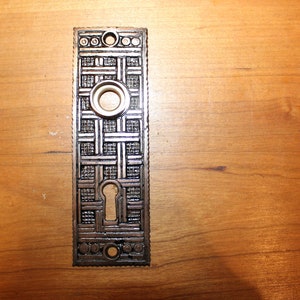 May include: A silver door plate with a geometric design and a keyhole. The plate has a decorative border with a series of circles.