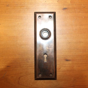 May include: A vintage, rectangular, brass door plate with a keyhole and a round knob. The plate has a dark patina and is mounted on a wooden surface.