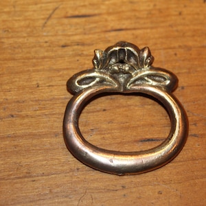 May include: A gold-toned metal ring pull with a decorative floral design. The ring pull is oval shaped and has a smooth surface.