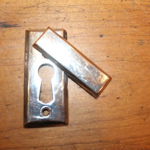 May include: A silver-toned metal door lock with a keyhole and a hinged cover. The cover is open, revealing the keyhole.