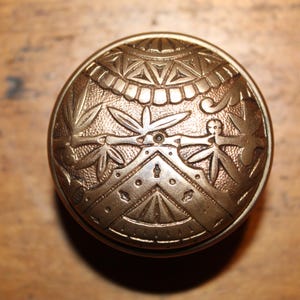 May include: A detailed, antique brass button with an intricate, embossed design. The button features geometric patterns, stylized leaves, and human-like figures. The button is circular and has a slightly domed shape.