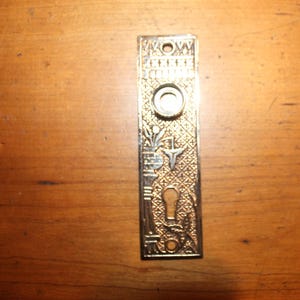 May include: Ornate, rectangular brass door plate with a keyhole and a circular knob. The plate features detailed floral and geometric patterns, with a vintage aesthetic. The brass has a warm, golden hue, suggesting an antique or vintage item.
