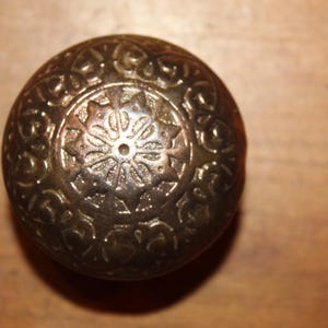May include: A round, brass door knob with an intricate, ornate design. The knob features a central floral motif surrounded by a repeating pattern.