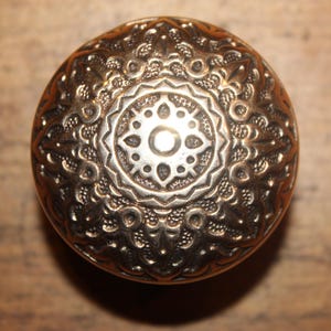 May include: A round, brass door knob with an intricate, ornate design. The knob features a central circular motif with a smaller circle in the center.