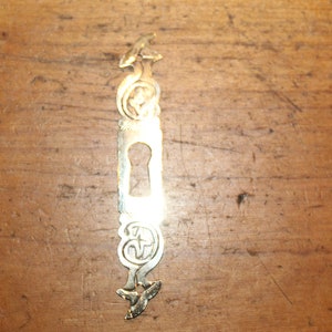 May include: A gold-colored metal keyhole cover with an ornate design. The cover has a keyhole in the center and decorative flourishes at the top and bottom.